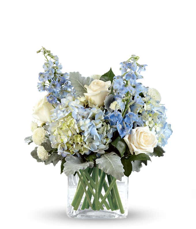 White roses, blue hydrangea, blue delphinium, and fresh greens arranged into a short cube vase