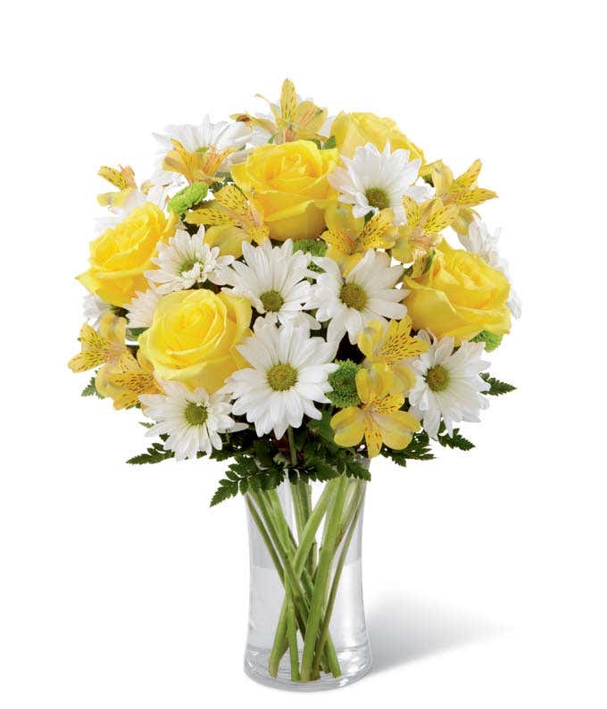 Bright bouquet featuring yellow roses, yellow alstroemeria, white daisies, green poms, and assorted greenery arranged in a tall clear glass vase. 