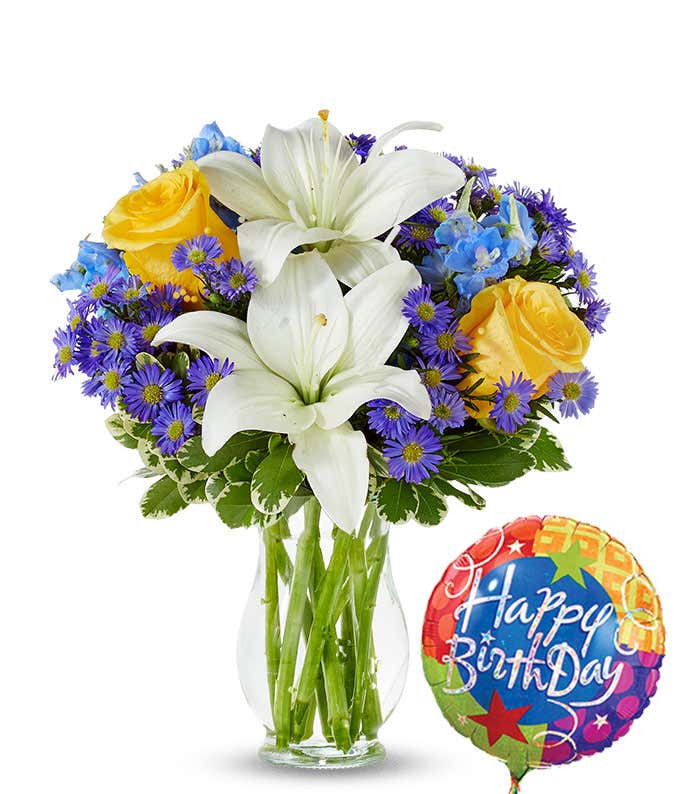 Birthday flower bouquet with white lilies, yellow roses, blue delphinium, and purple asters in a clear glass vase, paired with a colorful Happy Birthday balloon.