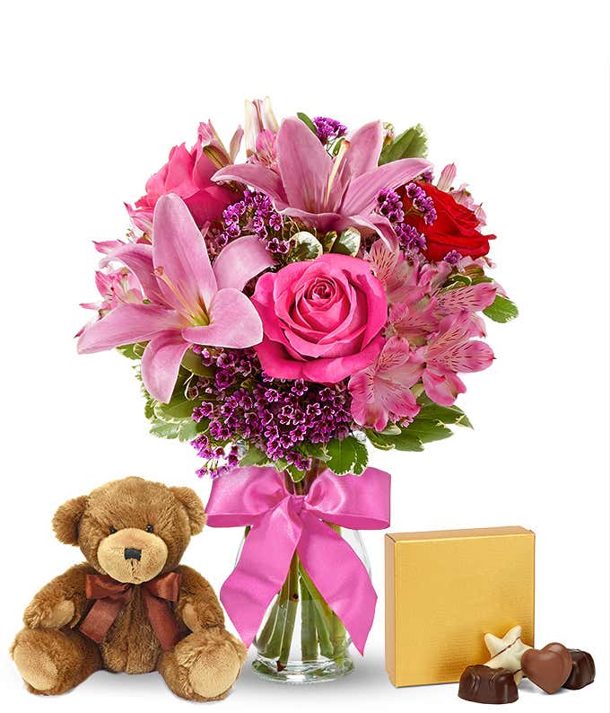 Romantic gift set with a bouquet of pink lilies, hot pink and red roses, pink alstroemeria, and purple filler in a glass vase with a pink bow, paired with a plush teddy bear and box of assorted chocolates. 