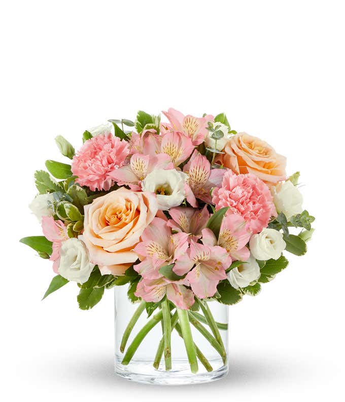 Bouquet of peach roses, pink carnations, pink alstroemeria, and white lisianthus with greenery, arranged in a clear glass cylinder vase.