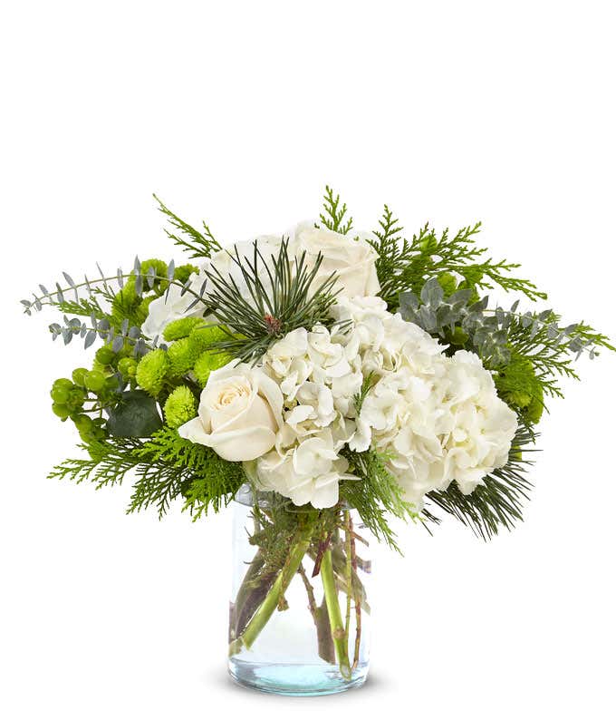 Bouquet of white hydrangeas, white roses, green button poms, green hypericum berries, and assorted evergreen foliage arranged in a clear glass cylinder vase.