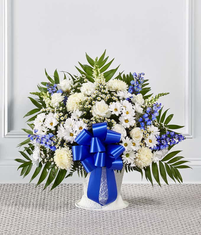 Standing floral arrangement of white roses, chrysanthemums, carnations, daisies, and snapdragons accented with blue delphinium and a large royal blue ribbon bow.