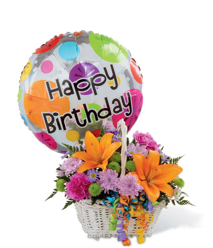 Festive birthday flower arrangement with orange lilies, pink carnations, purple poms, green button poms, and a colorful “Happy Birthday” balloon in a white basket.