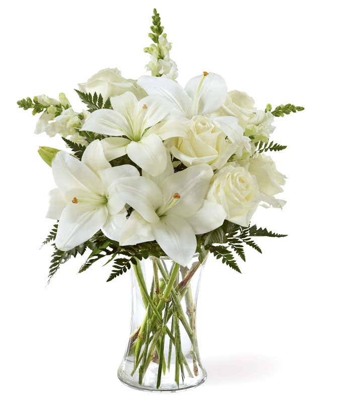 A bouquet of white lilies, roses, and snapdragons, with fresh floral greens, in a tall clear cylinder vase