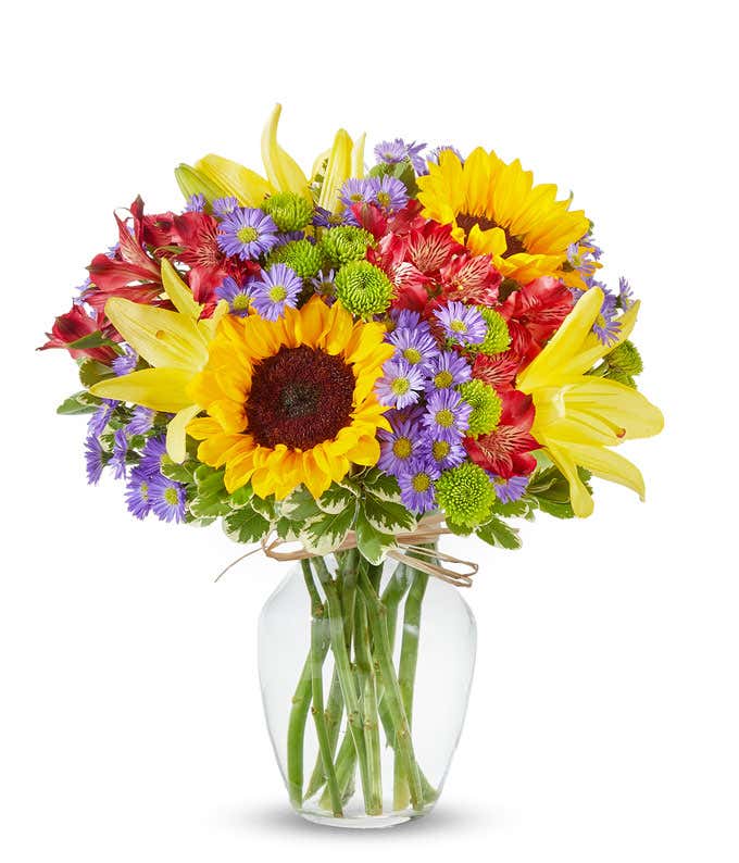 An arrangement of sunflowers, yellow lilies, orange roses, alstroemeria, Monte Casino, and green poms, in a clear glass vase.