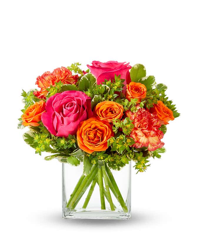 An arrangement of hot pink roses, orange spray roses, orange carnations, bupleurum, lily grass, and pittosporum, all beautifully arranged in a clear glass cube vase.
