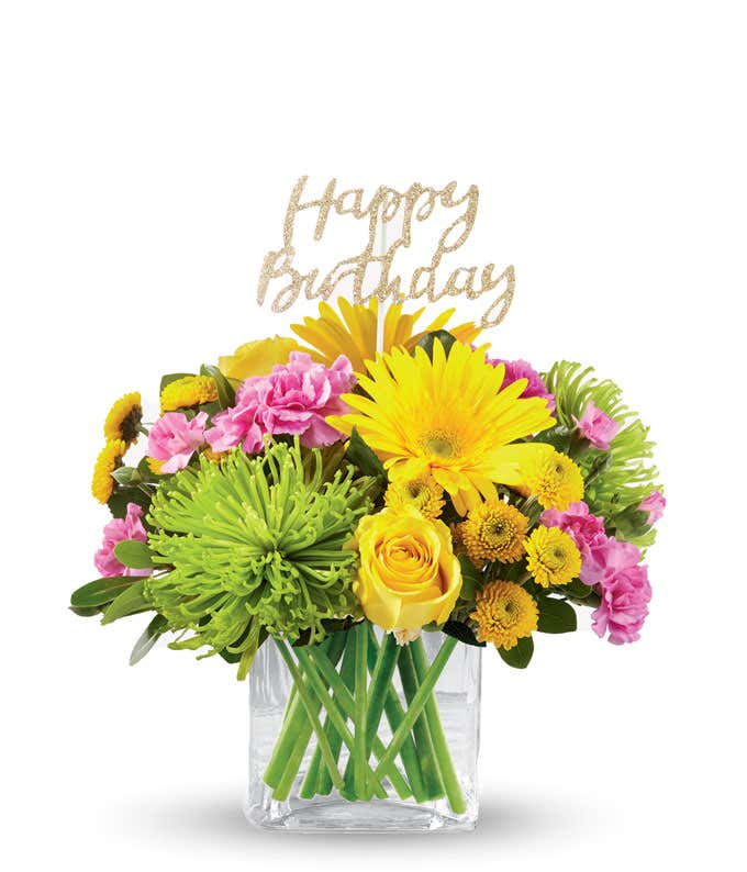 Yellow daisies and roes, pink carns, and green mums with a gold happy birthday pick, in a clear cube vase
