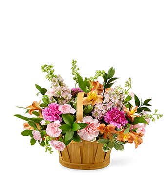 A floral arrangement featuring pink and coral roses (deluxe and premium sizes), pink carnations, pink mini carnations, orange alstroemeria, and pink larkspur, arranged in a wooden basket.