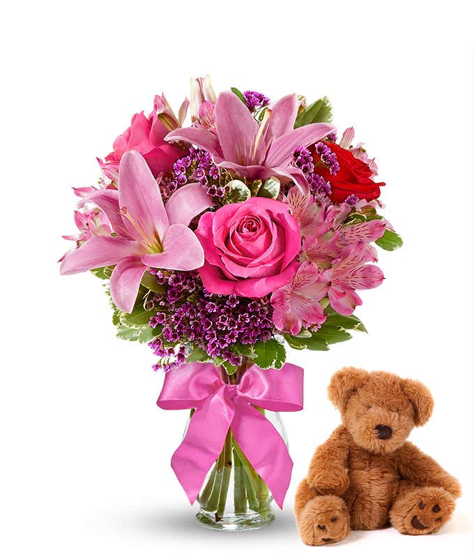 Red and pink roses, pink alstroemeria in a clear vase with bow, accompanied by a keepsake teddy bear.