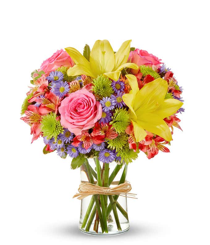 A vibrant arrangement featuring yellow lilies, pink roses, pink alstroemeria, green poms, and purple Monte Casino, beautifully arranged in a glass vase.