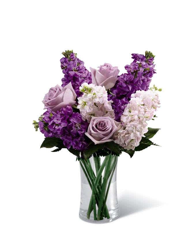 A bouquet of lavender roses and light and dark purple stock arranged in a clear glass vase with green leaves.