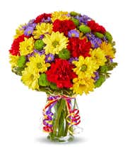 A bouquet of yellow daisies, red carnations, green poms, and purple asters in a clear vase with ribbon curls.