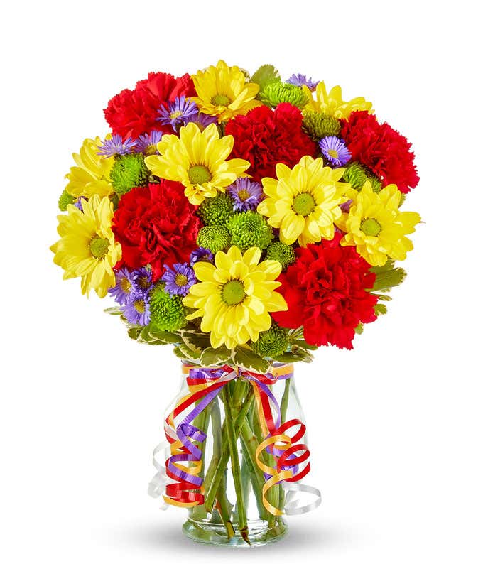 A bouquet of yellow daisies, red carnations, green poms, and purple asters in a clear vase with ribbon curls.