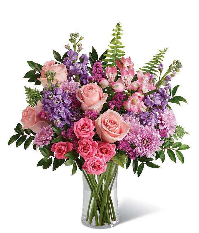 A bouquet of pink roses, lavender stock, purple mums, alstroemeria, and greenery in a clear glass vase.