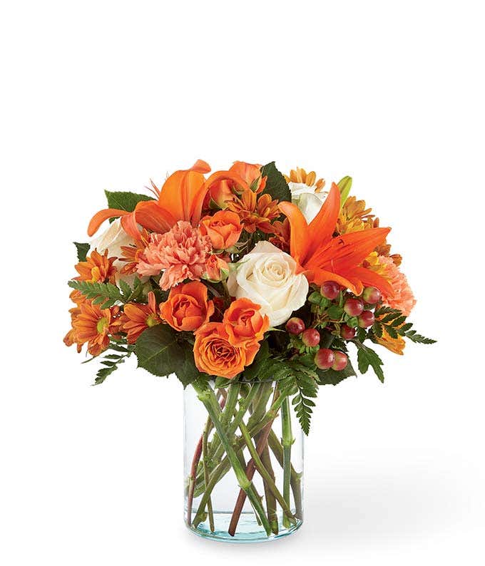 Clear glass vase filled with orange lilies, white roses, orange spray roses, peach carnations, orange daisy poms, red hypericum berries, and greenery.
