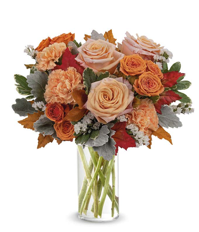 Clear glass vase filled with a lush arrangement of peach and orange roses, orange carnations, white filler flowers, dusty miller, red and orange autumn leaves, and assorted greenery.