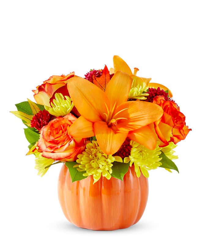 Bright fall flower arrangement with orange lilies, orange roses, yellow poms, red accent blooms, and greenery in a ceramic pumpkin container.