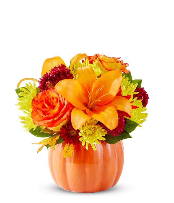 Bright fall flower arrangement with orange lilies, orange roses, yellow poms, red accent blooms, and greenery in a ceramic pumpkin container.