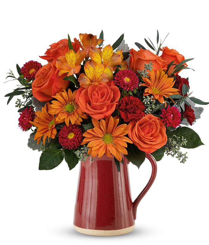 Orange Roses, Orange Alstroemeria, Bronze Daisies, Red Asters, Mini Maroon Carnations, and floral greenery in a red ceramic pitcher