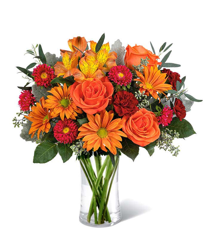 A vibrant floral arrangement featuring orange roses, orange alstroemeria, bronze daisies, red Matsumoto asters, maroon mini carnations, and seasonal greenery, complemented by a personal card message, all beautifully presented in a clear cylinder vase.