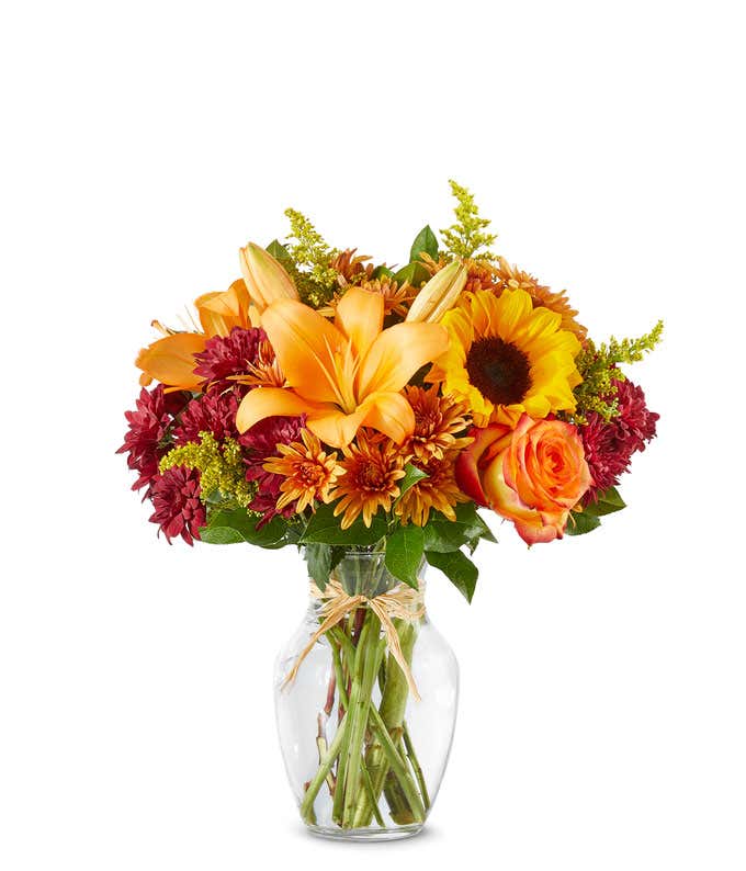 Bright autumn bouquet with sunflowers, orange lilies, orange roses, burgundy poms, yellow solidago, and butterscotch daisies in a clear glass vase tied with a raffia bow.