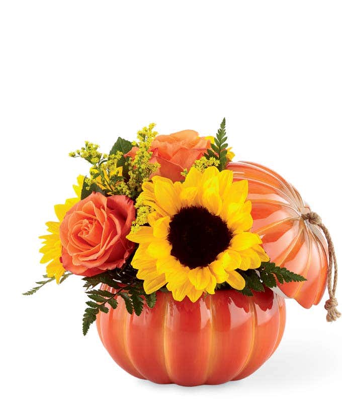 Fall flower arrangement with orange roses, sunflowers, yellow solidago, peach hypericum berries, and greenery in a ceramic pumpkin container with a lid and rope accent.