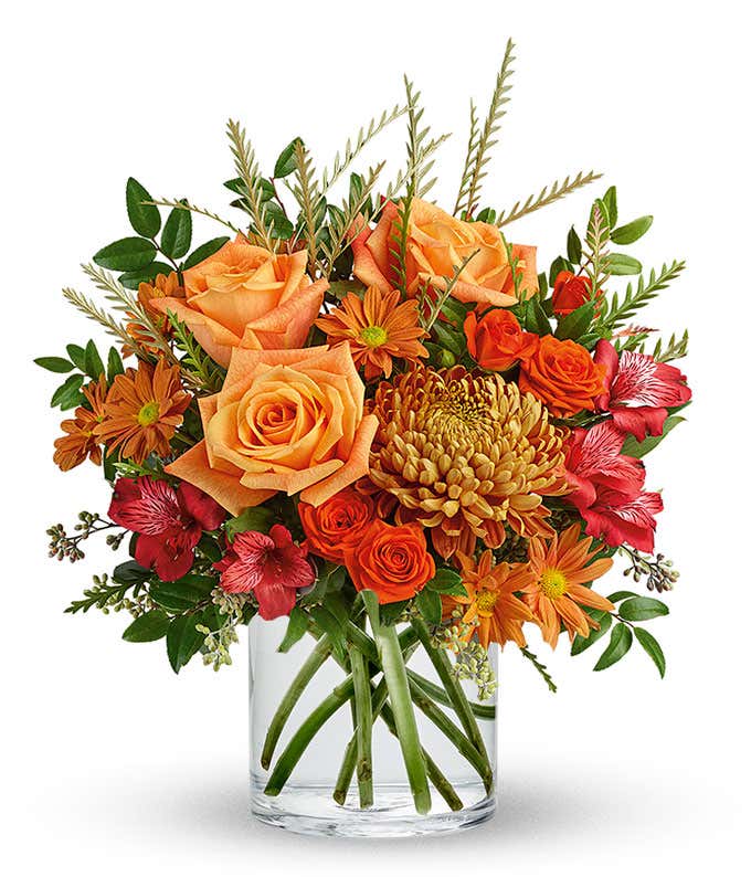 Autumn floral arrangement with orange roses, bronze daisy poms, red alstroemeria, golden mum, orange spray roses, and greenery in a clear glass cylinder vase.