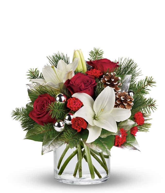 A festive arrangement with red roses, white Asiatic lilies, red mini-carnations, seasonal greens, silver ornaments, and pinecones, beautifully displayed in a clear cylinder vase.