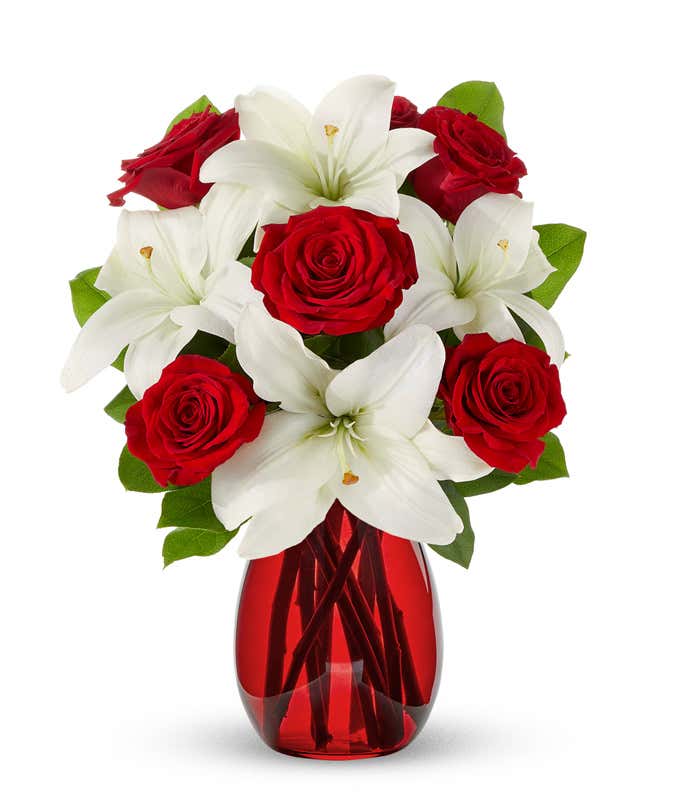 A floral arrangement featuring red roses and white lilies in a red cylinder vase.