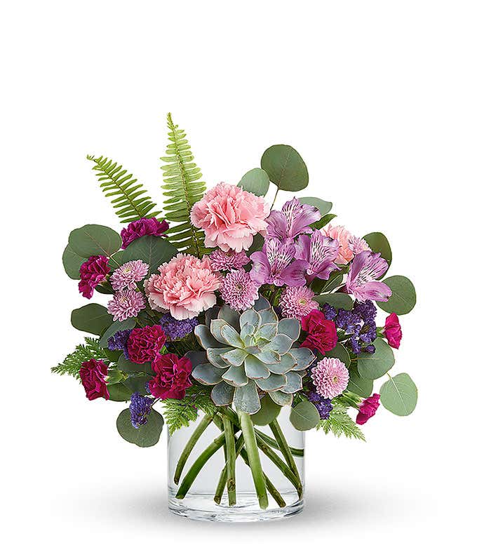 Floral arrangement featuring a succulent plant, pink carnations, purple mini carnations, dark pink roses (in deluxe and premium versions), purple statice, purple alstroemeria, lavender button mums, seasonal greenery in a glass vase