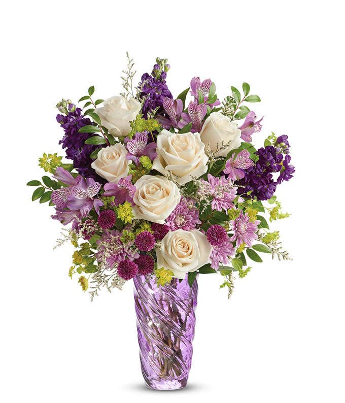 Floral arrangement featuring crème roses, lavender alstroemeria, purple stock, purple button chrysanthemums, lavender cushion spray chrysanthemums, white limonium, bupleurum, and huckleberry, all elegantly arranged in a lavender swirled vase.