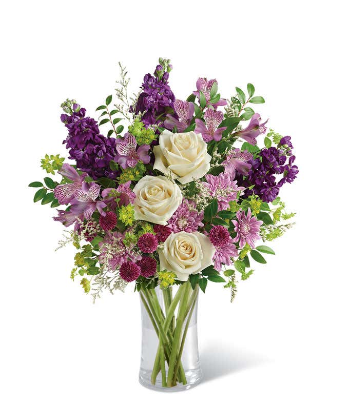A sophisticated floral arrangement featuring crème roses, lavender alstroemeria, purple stock, purple button chrysanthemums, lavender cushion spray chrysanthemums, white limonium, bupleurum, and huckleberry, all elegantly arranged in a cylinder vase.