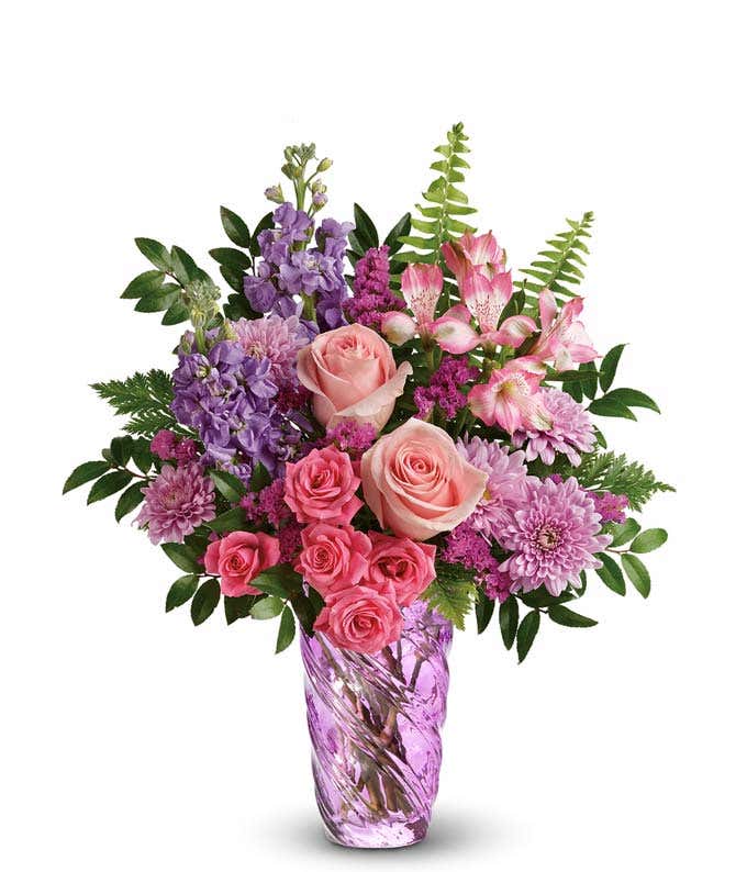 Full bouquet of pink roses, hot pink spray roses, lavender stock, pink alstroemeria, purple poms, and greenery arranged in a lavender swirl glass vase.