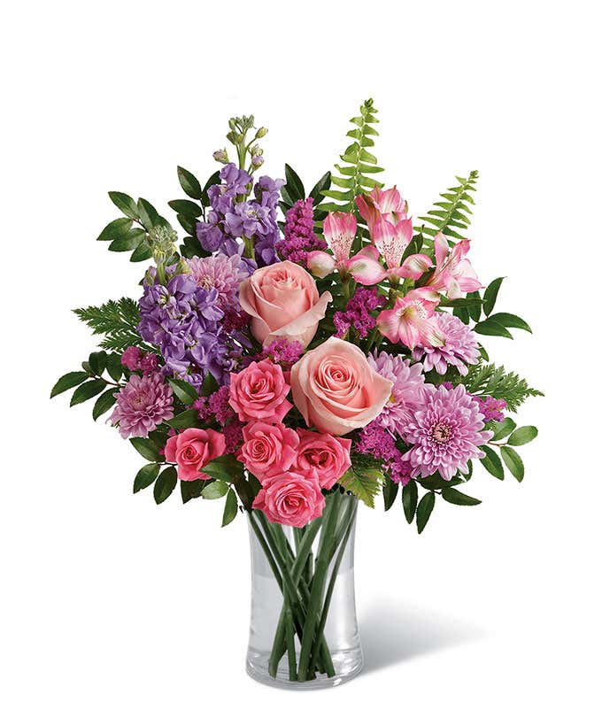 Floral arrangement featuring light pink roses, pink spray roses, pink alstroemeria, lavender stock, lavender mums, raspberry statice, and greenery in a glass vase.