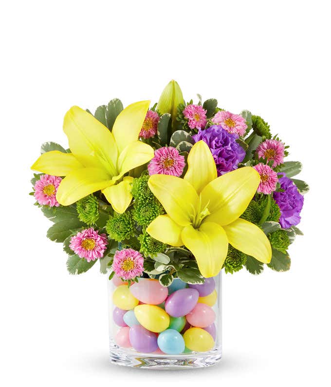 Yellow Asiatic lilies, yellow roses, purple carnations, pink Matsumoto asters, and green button poms arranged in a clear glass vase filled with colorful pastel Easter eggs