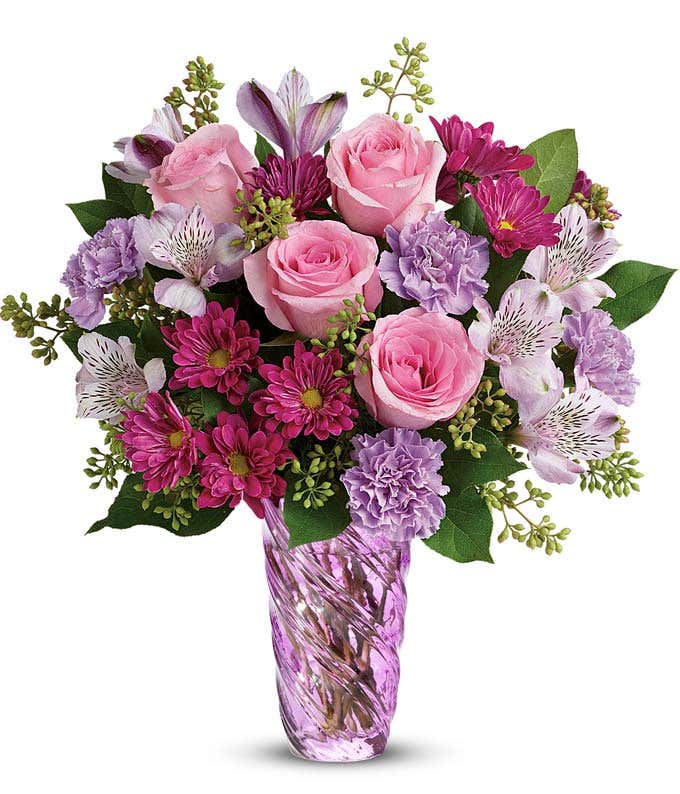 A bouquet of pink roses, lavender alstroemeria and carnations, purple daisies, with seeded eucalyptus exploding out of a swirled lavender cylinder vase.