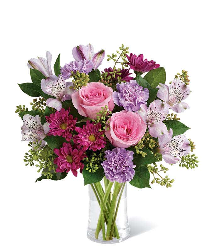 Pink roses, lavender carnations, magenta daisies, white alstroemeria, and lush greenery arranged in a clear glass vase, creating a soft and elegant bouquet in shades of pink and purple.