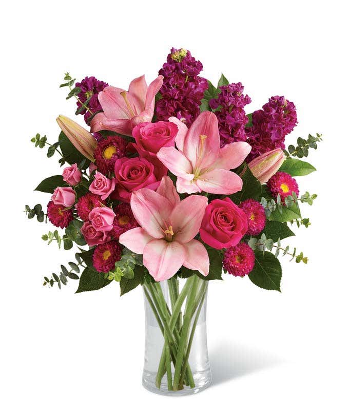 Pink lilies, hot pink roses, pink spray roses, magenta Matsumoto asters, and purple stock arranged with eucalyptus and greenery in a clear glass vase, creating a bold and elegant pink floral bouquet.