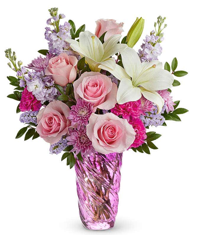 Bouquet of pink roses, white lilies, pink carnations, lavender stock, and purple poms arranged in a twisted lavender glass vase with greenery.