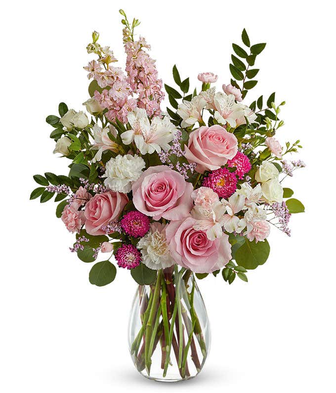 arrangement featuring Pink Roses, White Spray Roses, White Carnations, Ivory Alstroemeria, Miniature Pink Carnations, Pink Larkspur, Hot Pink Matsumoto Asters, and floral greens, all beautifully presented in a clear glass vase