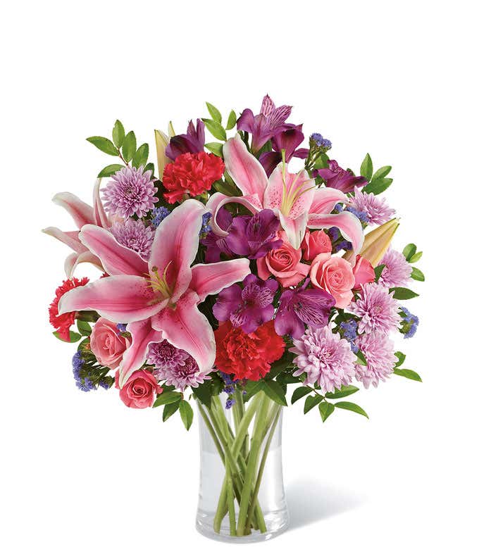 A vibrant bouquet of fresh pink lilies, red carnations, lavender chrysanthemums, purple alstroemeria, pink roses, and greenery arranged in a clear glass vase. The lush mix of colors creates a cheerful and elegant floral display.