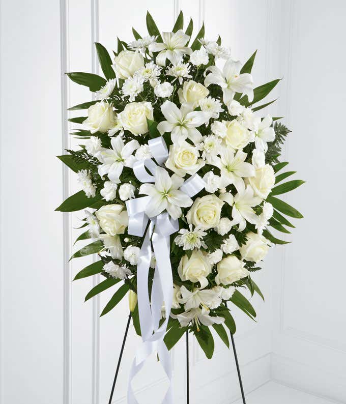 A standing spray made up of white lilies, roses, mums, and carnations, with fresh floral greens and a white ribbon