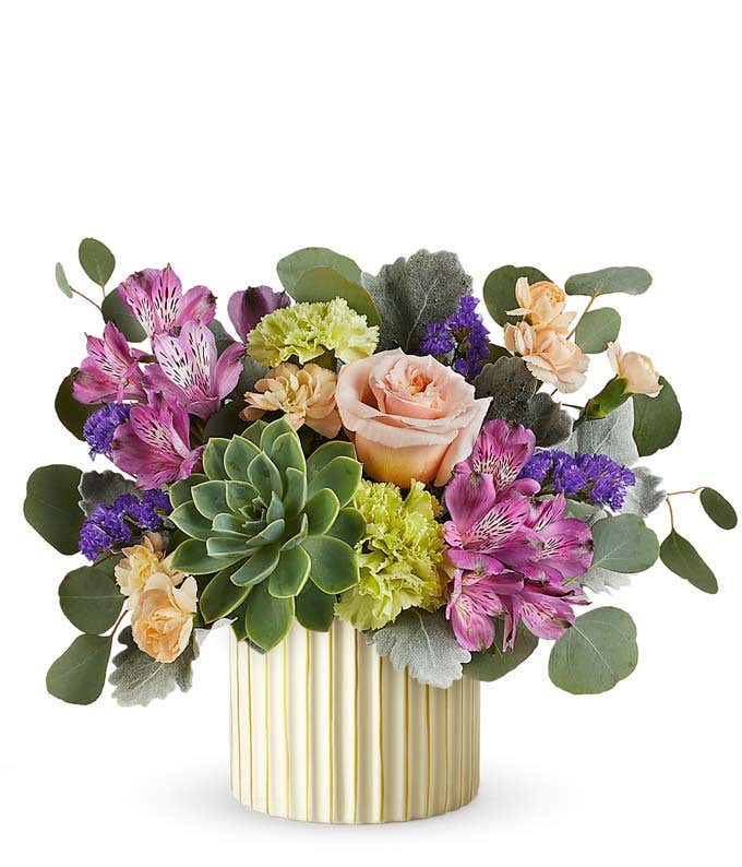 Floral arrangement with blush roses, purple alstroemeria, green carnations, small peach blooms, a large green succulent, and eucalyptus in a ribbed ivory planter with gold accents.