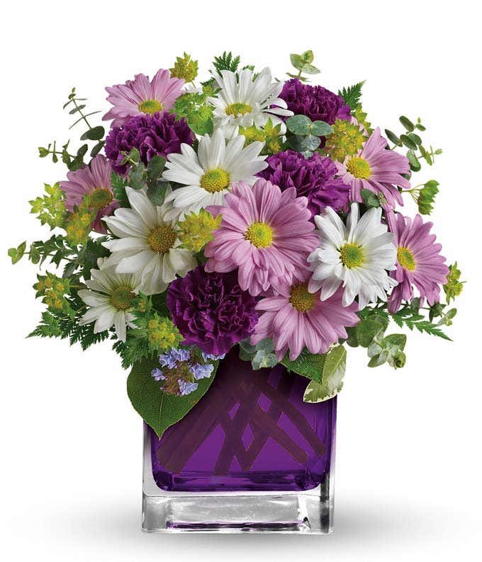 A beautiful arrangement featuring purple carnations, lavender daisy spray mums, white daisy spray mums, bupleurum, spiral eucalyptus, and leatherleaf fern, presented in a purple glass cube vase.