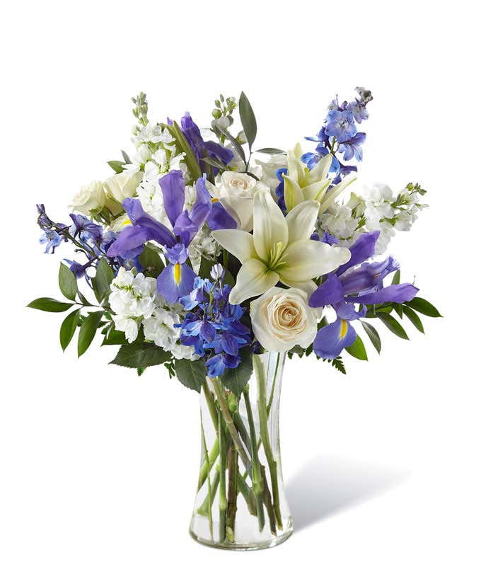 White lilies, blue delphinium, and white roses with fresh floral greens arranged into a tall clear cylinder vase