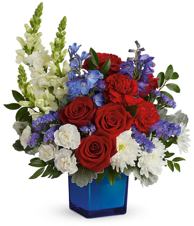 Memorial Day arrangement featuring red roses, red carnations, white miniature carnations, blue delphinium, white snapdragons, white cushion chrysanthemums, and lush floral greenery in a blue glass cube vase.