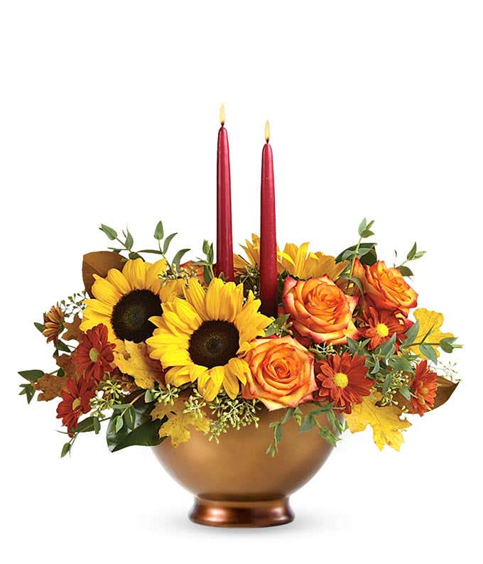 Centerpiece with Orange Roses, Orange Asiatic Lilies, Maroon Mini Carnations, Orange Cushion Spray Mums, and floral greenery with burgundy tapered candles in a bronze bowl