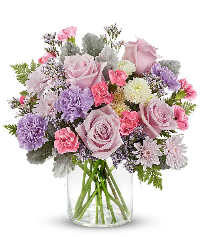 Soft pastel floral arrangement featuring lavender roses, lavender carnations, pink miniature carnations, lavender cushion spray chrysanthemums, white button spray chrysanthemums, dusty miller, and lush greenery in a short cylinder vase.