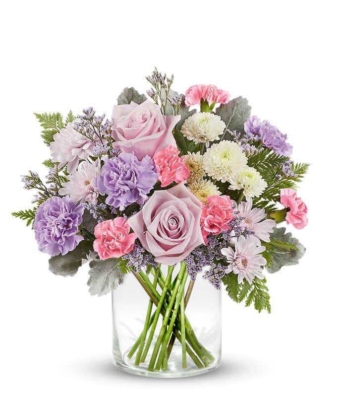 Soft pastel floral arrangement featuring lavender roses, lavender carnations, pink miniature carnations, lavender cushion spray chrysanthemums, white button spray chrysanthemums, dusty miller, and lush greenery in a short cylinder vase.